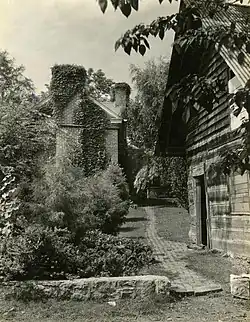 Black & White photo showing rough-stone knee-wall in foreground; mansion house in back; and a log building to the right; all interspersed with foliage