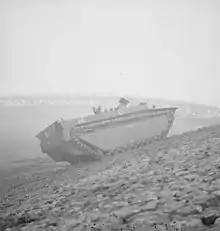 A tracked armoured vehicle drives up the bank of a river. In the background lies the wrecked frame of a metal bridge.