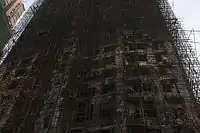 An upward view of a burned-out high-rise: a blackened mess of tangled scaffolding and holes for windows.