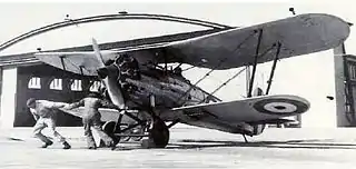 Two men swinging the propeller of a biplane in front of a hangar