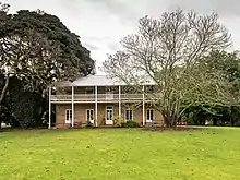 Two storey sandstone homestead stands on a grassy area surrounded by large trees