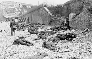 The deadly result of enfilade fire during the Dieppe Raid of 1942: dead Canadian soldiers lie where they fell. Trapped between the beach and fortified sea wall (covered with barbed wire), they made easy targets for MG 34 machineguns in a German bunker. The bunker firing slit is visible in the distance, just above the German soldier's head.