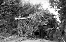 Three men conduct maintenance on a tank; the tank is partially obscured by bushes, which its gun barrel protrudes through.