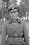 A man wearing a military uniform and coat, peaked cap and a neck order in the shape of a cross. His cap has an emblem in shape of a human skull and crossed bones.