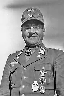 A man wearing a field cap and military uniform with various military decorations including an Iron Cross displayed at the front of his uniform collar.