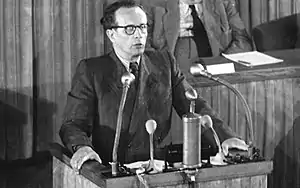 A man with glasses talking into microphones behind a lecturn.