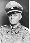 A man wearing a military uniform, peaked cap and a neck order in the shape of a cross. His cap has an emblem in shape of a human skull and crossed bones.