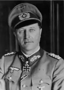 The head and shoulders of a man. He wears a peaked cap and military uniform, and an Iron Cross displayed at the front of his uniform collar. His facial expression is a determined; his eyes are looking into the camera.