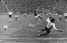 A black-and-white photograph of footballers in action, taken from behind the goal as a penalty kick is taken. The ball has been kicked by the taker to the left of the goalkeeper, who has dived forlornly to his right.