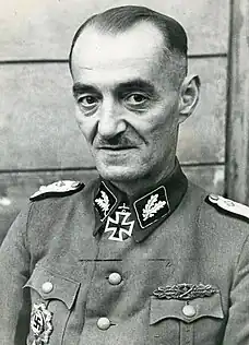 A man wearing a military uniform. He has short, thinning hair, mustache and a determined facial expression.