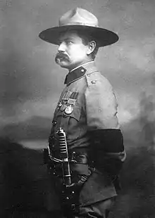 Photo of Burnham taken in 1901 in London after his investiture with the cross of the Distinguished Service Order by King Edward VII.  He is dressed in British Army uniform and standing at attention, facing right.  On his left arm is a black armband worn in mourning for the recent death of Queen Victoria. He is wearing his stetson hat and a sword on his left side. He sports a large handlebar mustache.