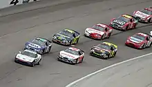 a single road car leads a group of race cars which are being driven in a double-file queue
