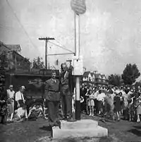 A man in a suit standing on a stage next to a large pole. There is a Marine in the background in his dress uniform and behind him a crowd of people are watching the man on the stage.