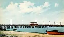 An image of a red streetcar on a wooden trestle, crossing a body of water. A beach with several small boats is visible in the foreground.