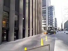 The recessed plaza surrounding the CBS Building, looking west along 53rd Street. The plaza has gray pavement and is several steps below the street. There are yellow cones in the plaza. To the left is the CBS Building, and to the right is the sidewalk on 53rd Street.