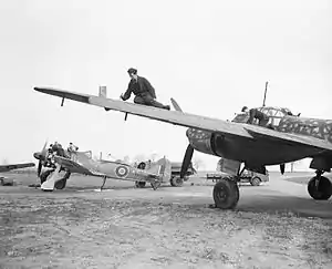 A photograph of a single–engined and a larger, twin-engined aircraft on the ground. Two men are working on the wing of the larger aircraft. The engine–covers have been removed from the smaller aircraft and four men are working on its engine