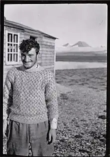 Brian harland is standing outside a hut in Svalbard, 1938. He is wearing a jumper and smiling.