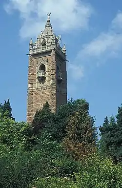 An ornate brick tower surrounded by trees. The tower has balconies and is surmounted by a pitched roof with an ornate figure at the apex.