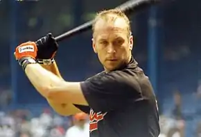 Cal Ripken Jr. in an Orioles shirt, wearing orange batting gloves, and no hat. He's holding the bat as he prepares to take a practice swing, looking towards the camera.