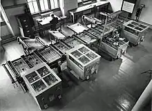 Looking down from above at a room at an intricate mechanical device which fills the room. In the background, a man sits at a desk next to a filing cabinet. Owens 1991, p. 15.