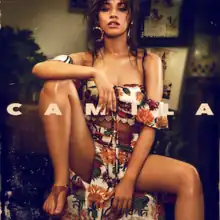 The cover image features Camila Cabello in foreground sitting on a small circular table wearing nature themed bra-like crop-top and long-skirt. In the background is a dim-lighted living-room. Above everything, in the middle, spread over whole width, in capitalized and bold format, is written the title 'CAMILA'.