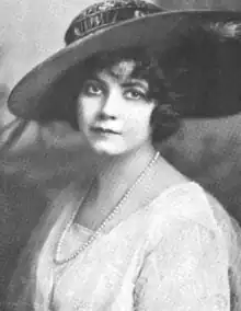 A young white woman with dark hair and eyes, wearing a wide-brimmed hat, a strand of pearls, and a white dress or blouse with a square neckline