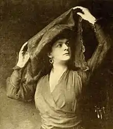 Black and white photograph of a dark-haired woman in a long-sleeved garment holding a piece of fabric above her head.