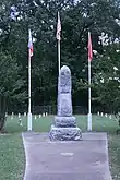 Photo of the 12-foot memorial stone obelisk erected in the center of the cemetery with three flagpoles in background containing flags of United States, Texas, and Arkansas