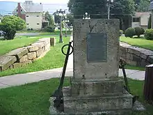 A stone and metal monument and stone retaining walls commemorate a former canal lock. A sidewalk runs by the memorial and between the retaining walls on a lush bed of grass.