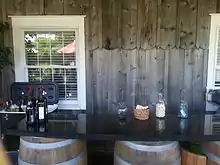 A black marble bar with wine bottles sitting atop oak barrels, with a wooden wall and white-colored window in the background.