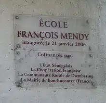 A clear plaque on a grey wall stating "École François Mendy" at the top with smaller writing below