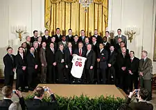 Three rows of men in various dark-colored suits; in the center, a gray-haired smiling man holds a white baseball jersey that reads "Bush" on the back in small red print with "06" in larger red print below it.