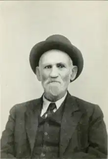 A man with a white beard, wearing a three-piece suit and a hat with a rounded brim faces the camera