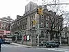 A three-story brick and stone building sits on a corner in New York City and is surrounded by a stone and wrought iron fence.