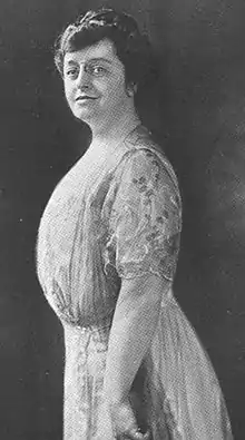 A middle-aged white woman standing almost in profile, wearing a short-sleeved light-colored gown. Her dark hair is arranged in an updo.
