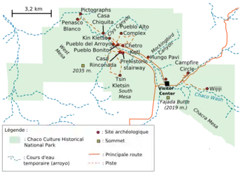 A large green area representing Chaco Culture National Historical Park's boundaries sits in the middle of a white field. The green area is roughly rectangular with one smaller square-like and one triangular appendage abutting it at bottom-left and bottom-right, respectively. Fifteen small red circles represent the location of important Chacoan sites; they are focused on a line running from top-left (northwest) to bottom-right (southeast). A dashed blue line depicting the Chaco Wash runs roughly along the same line; a network of dashed and solid orange lines represent trails and metalled roads, respectively, also focus on the same axis, connecting the red dots. Two gold squares define high points: "Fajada Butte (2019 m.)" and "West Mesa (2035 m.)".
