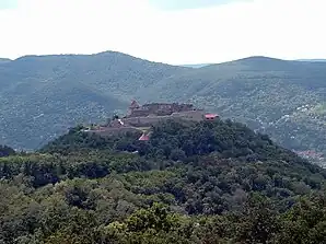 A castle atop a forested hill, surrounded by mountains.