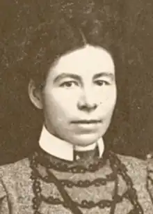 A young white woman with dark hair center parted and dressed up from the shoulders; wearing a high-collared dress with braid trim and puff sleeves