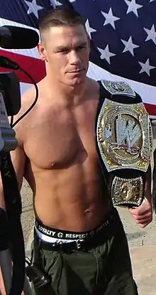 A man with a championship belt on his shoulder, with the American flag in the background.