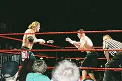 Two shirtless men with their arms held up in defensive positions stand crouched facing each other inside a wrestling ring with three red ropes running parallel to each other. A crowd of spectators watch from all side of the ring as a man wearing a referee shirt stands with his hands on his knees watching the two men.