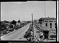 Center Street at 100 West facing west, with a parade on the south side of Center Street. Numerous automobiles line the street and businesses such as Hedquist Drugs, Wilkins Hotel, Taylor Brothers, and Smith Brothers Auto Repair Shop line the street