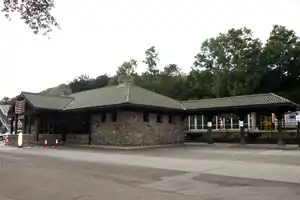 A brownish stone one-story building with a low hipped roof covered in green tile, seen from a three-quarter angle across pavement. From its right protrudes a similarly treated narrow pavilion; on its front, near the left side, is a small porte-cochere adorned with an American flag, its entrance blocked off by small orange plastic cones. Behind the structure are an iron fence, railroad tracks, some portions of modern concrete structures, and trees.