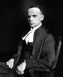 A dark-haired middle-aged man sits in a wooden chair, facing left. He is wearing judicial robes.