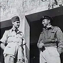 Two men in military uniform standing outside of a building talking to each other. The man on the left is wearing a lighter coloured uniform with short sleeves. The man on the right is wearing a long-sleeved, dark jacket.