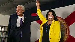 A man with white hair wearing a suit with a purple tie stands with a woman with black hair wearing a yellow jacket and a black shirt with a blue necklace.