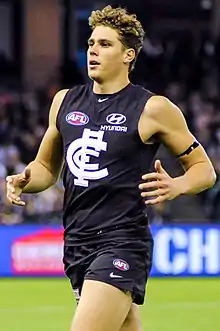 A man with short curly brown hair in a sleeveless navy guernsey and shorts