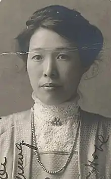 A young Asian woman wearing a high lace collared blouse, beads, and a jacket; her dark hair is in an updo. There is handwriting running over the photograph from the bottom edge.