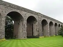 A series of seven stone archways, connected to stone parapet above grass area.