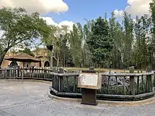 The Cheetah Hunt enclosure is depicted with parts of the ruined temple on the left side. A board is displayed in the foreground on the time of the Cheetah runs, with the enclosure in the background separated by a wooden fence. Tall trees surround the back side of the gated enclosure.