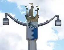 Post holding two lamps at the ends of two arms. The top of the post is decorated by a galleon; underneath the boat is a shield containing a white left and a blue right separated by a jagged line, on top of which rests a white bird carrying an olive branch.